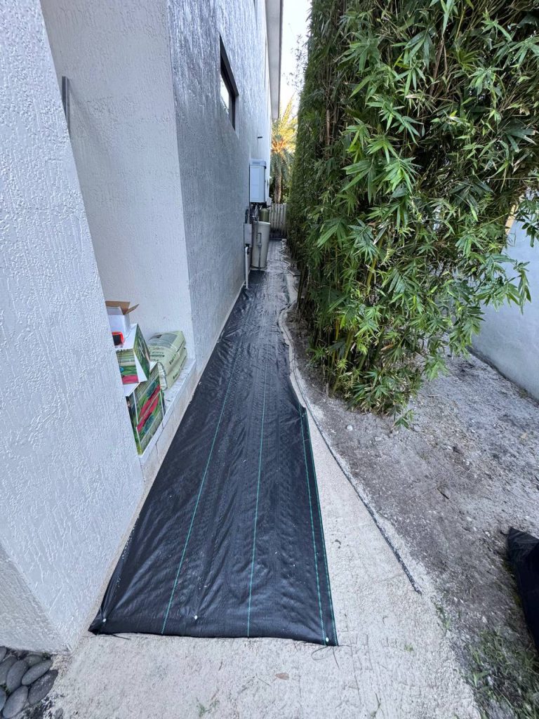 Dark installation underlay in a narrow passage between a white wall and bamboo.