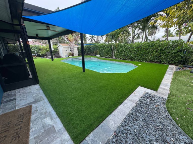 Turf around a swimming pool beneath a blue shade sail and dark posts.